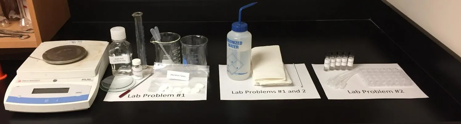 2017 USNCO Part III lab setup showing three zones: Problem 1 (balance, acetic acid, Alka-Seltzer tablets, glassware), shared equipment (deionized water), Problem 2 (unknown vials, well plate)