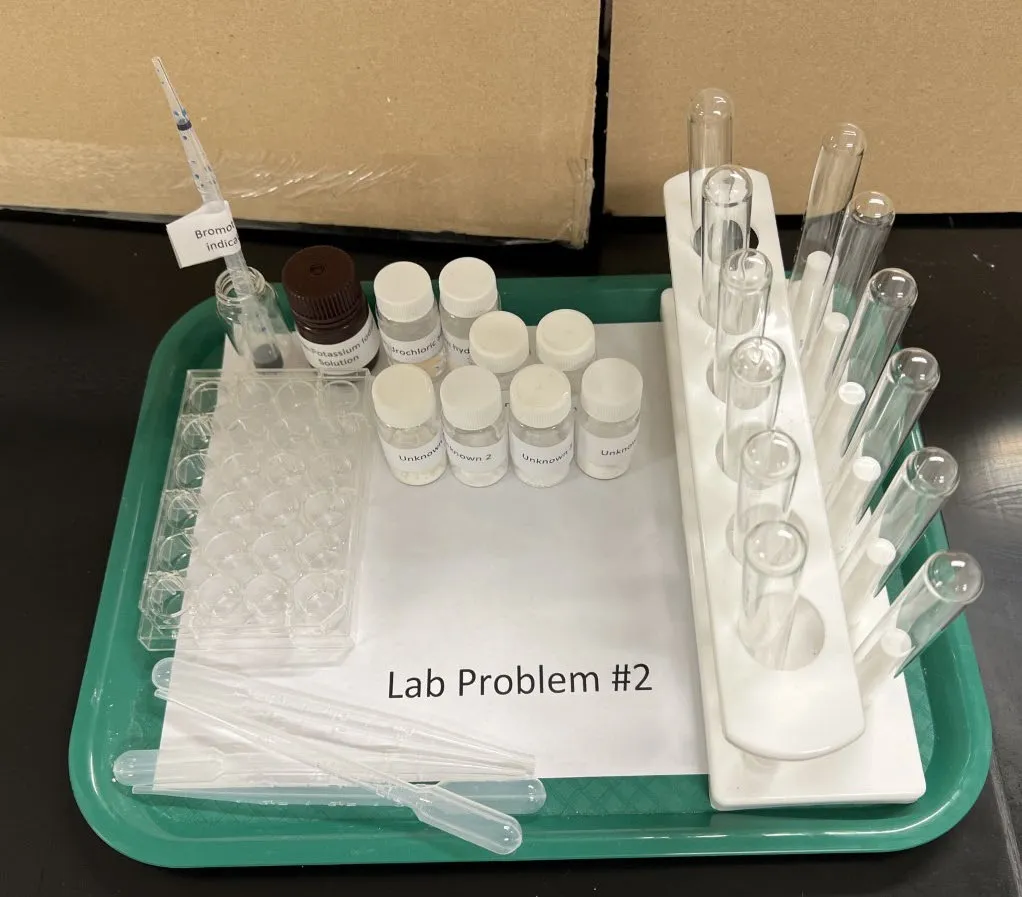 2022 USNCO Part III Problem 2 setup: unknown solution bottles, bromothymol blue indicator, test tubes in rack, well plate, Beral pipets on green tray